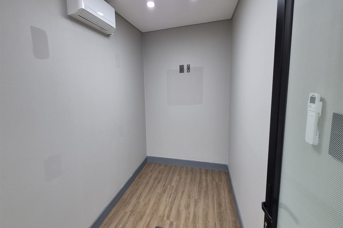 A compact, empty office space featuring laminate flooring, recessed lighting, a wall-mounted air conditioning unit, and glass-panelled entry door.