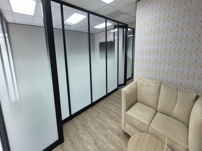 Modern office space featuring sleek black-framed glass partitions with frosted privacy film, wood-look flooring, and contemporary patterned wallpaper.