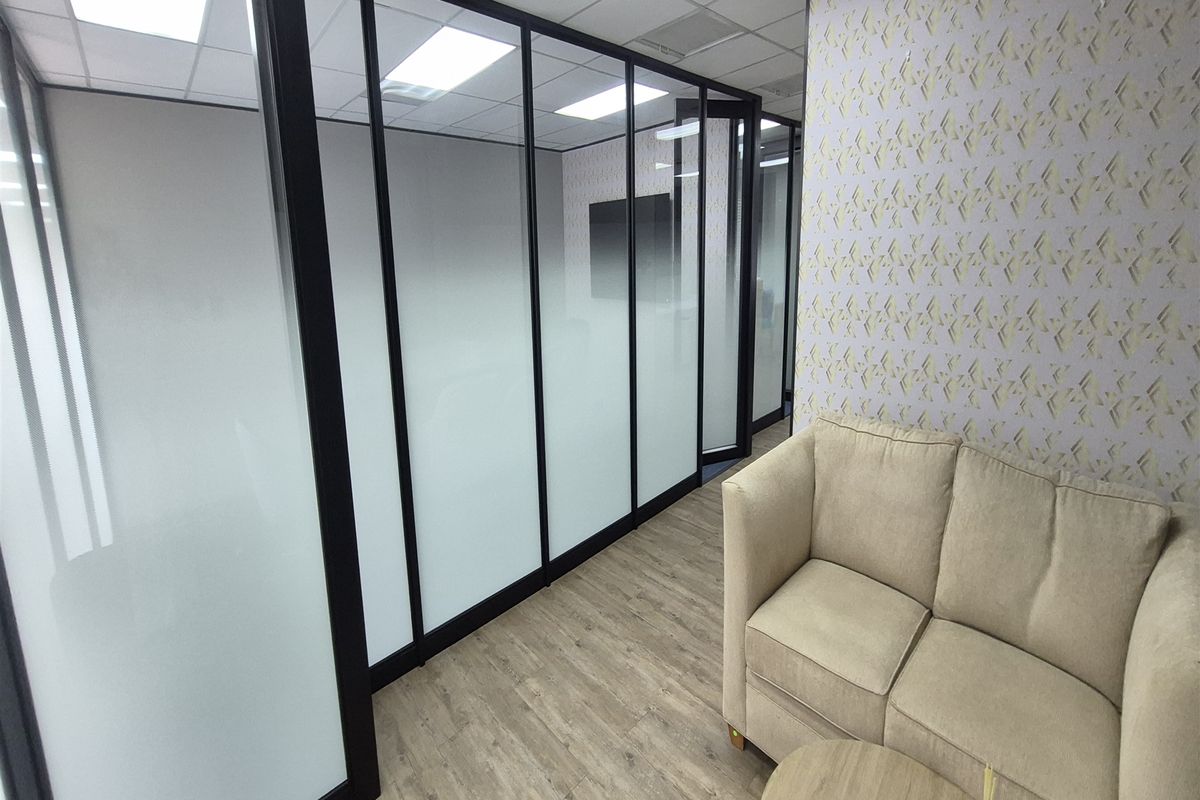 Modern office space featuring sleek black-framed glass partitions with frosted privacy film, wood-look flooring, and contemporary patterned wallpaper.