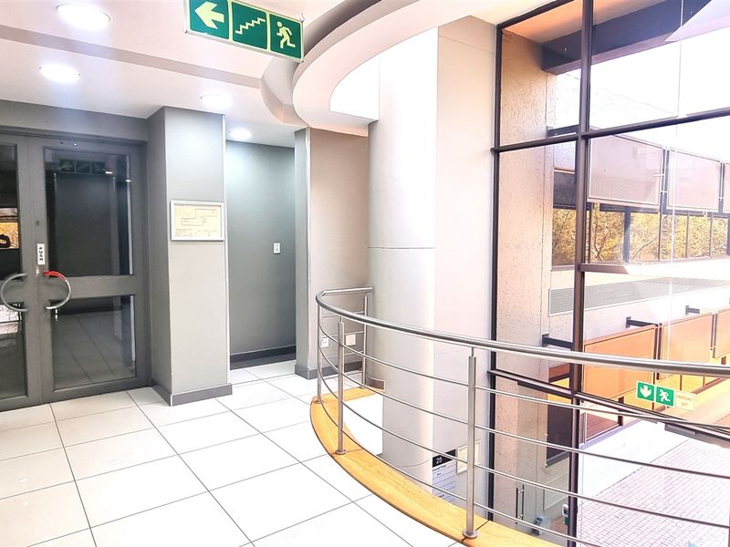Modern office building hallway featuring large glass curtain walls, stainless steel balustrades, and clean tiled flooring with recessed lighting.