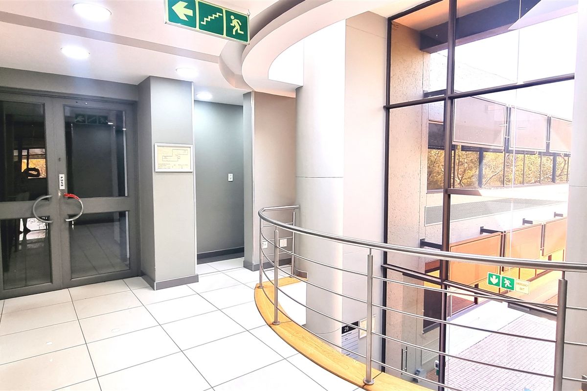Modern office building hallway featuring large glass curtain walls, stainless steel balustrades, and clean tiled flooring with recessed lighting.