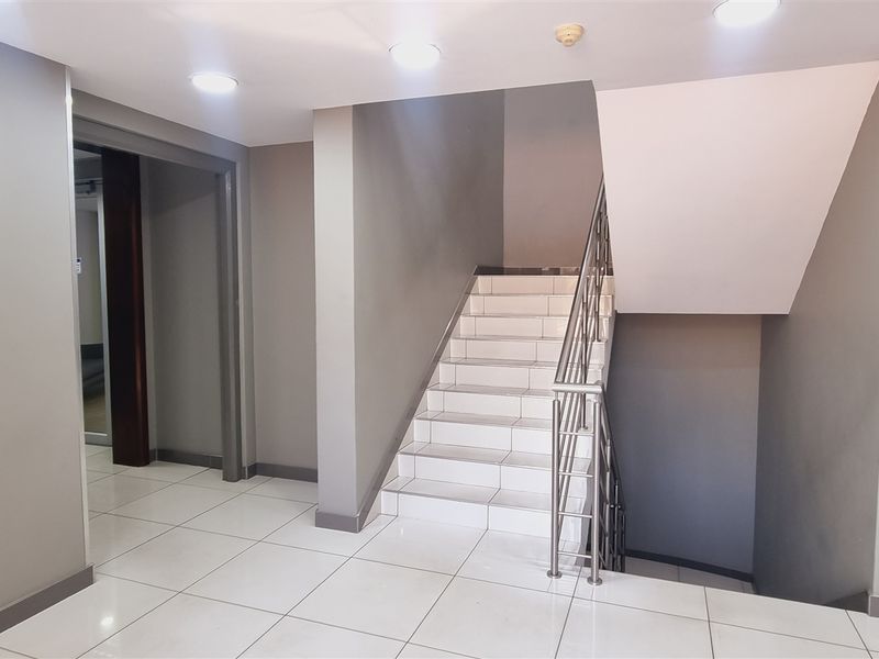 A clean, modern office hallway featuring white tiled flooring, a staircase with a stainless steel handrail, and recessed ceiling lighting.