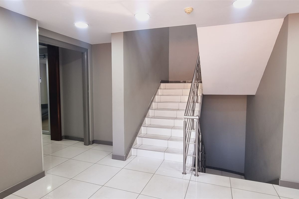 A clean, modern office hallway featuring white tiled flooring, a staircase with a stainless steel handrail, and recessed ceiling lighting.