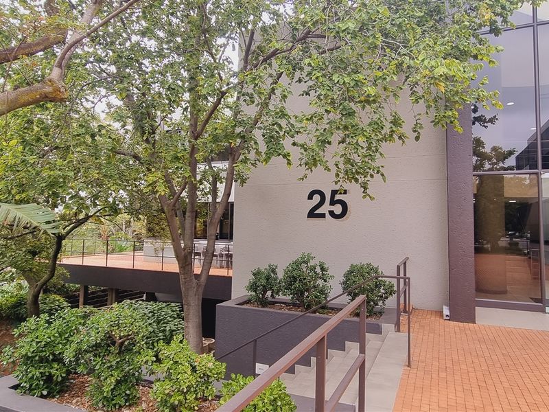 Modern office building entrance featuring a glass facade, brick paved walkway, and elevated deck area with mature landscaping.