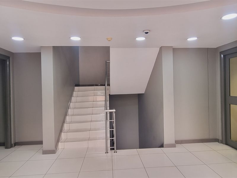 A clean, modern office hallway featuring white tiled flooring, recessed ceiling lighting, and a central staircase with a metal handrail.