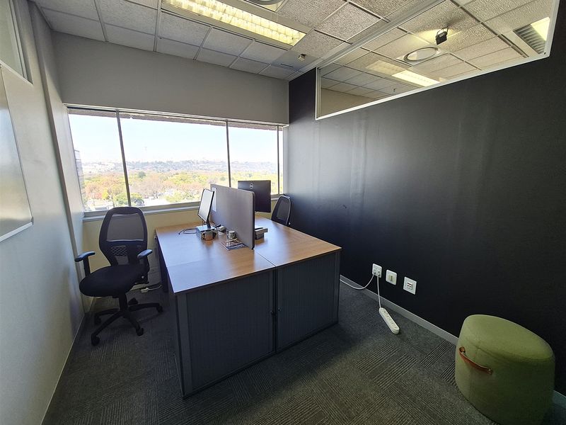 A professional office space featuring carpeted flooring, a suspended ceiling with recessed lighting, and a large window providing natural light and exterior views.