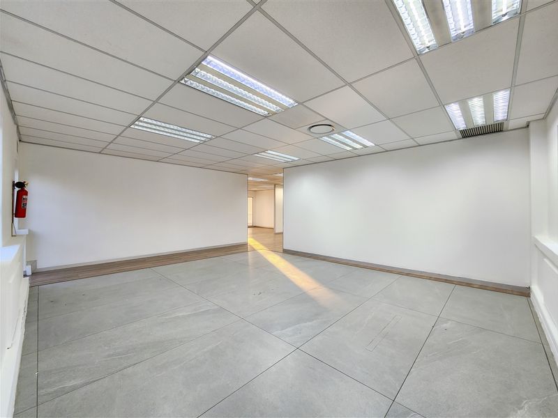 A bright, vacant office space featuring neutral tiled flooring, a suspended ceiling with recessed lighting, and a wall-mounted fire extinguisher.
