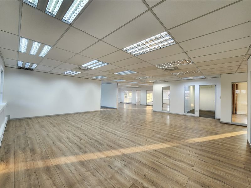 A spacious, open-plan office area featuring modern laminate wood flooring, a suspended ceiling with recessed lighting, and glass partition doors.