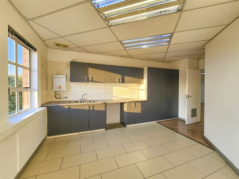 A functional office kitchenette featuring dark grey cabinetry, laminate countertops, a stainless steel sink, and tiled flooring under a suspended ceiling.