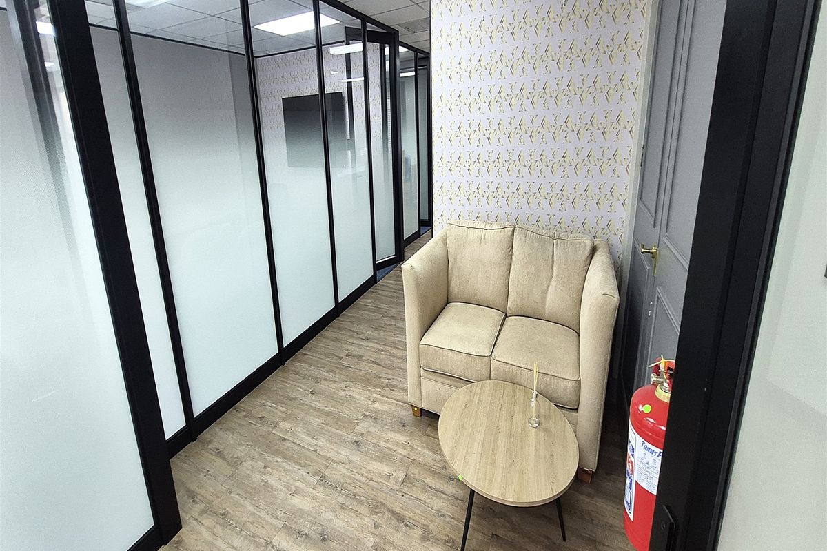 A modern office space featuring glass partition walls, wood-look laminate flooring, and a suspended ceiling with recessed lighting.
