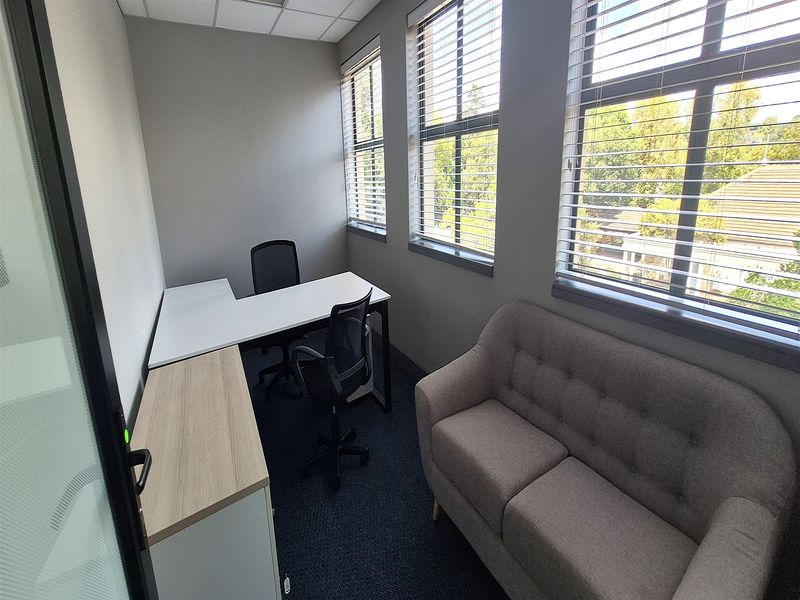 A compact, well-lit private office space featuring carpeted flooring, suspended ceiling, and large windows with venetian blinds providing natural light.