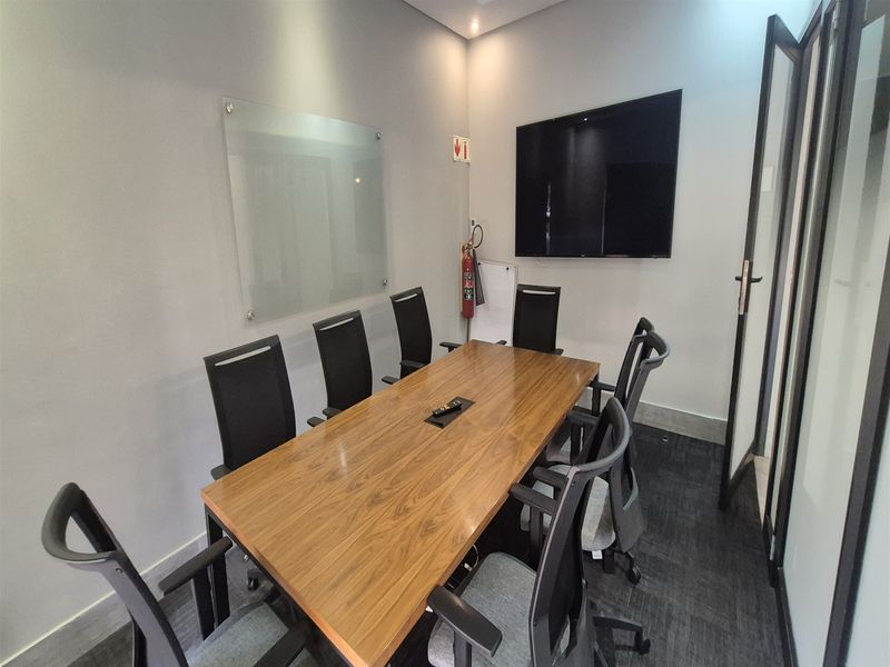 A professional meeting room featuring a wooden conference table, glass whiteboard, wall-mounted display, and modern glass partition entry.
