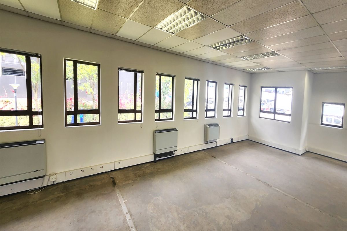 An empty office space featuring a suspended ceiling, recessed lighting, wall-mounted HVAC units, and multiple windows with perimeter electrical trunking.