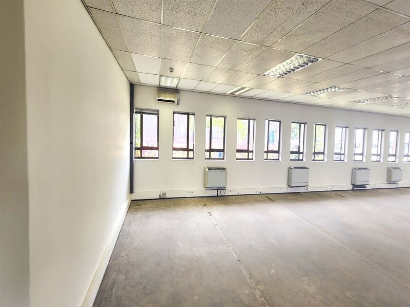 An empty, open-plan office space featuring a suspended ceiling, recessed lighting, multiple windows, and wall-mounted air conditioning units with exposed concrete flooring.