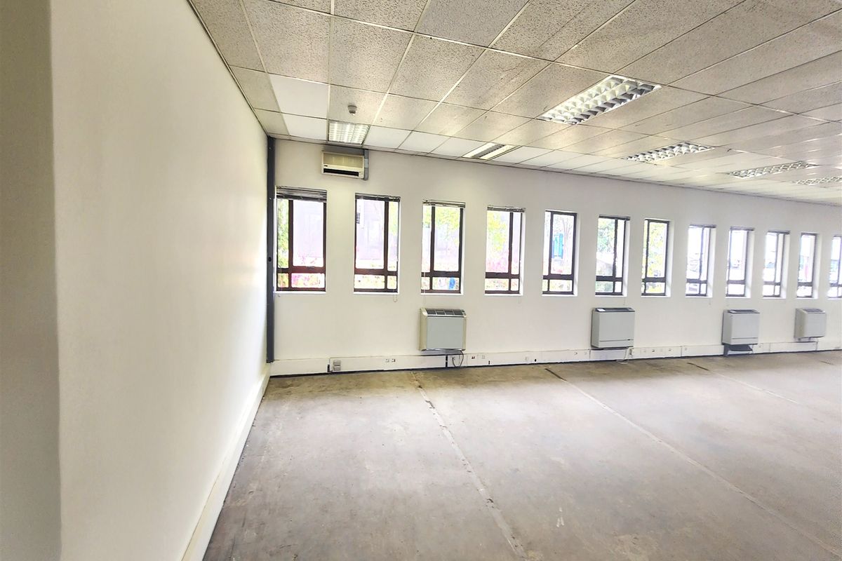 An empty, open-plan office space featuring a suspended ceiling, recessed lighting, multiple windows, and wall-mounted air conditioning units with exposed concrete flooring.
