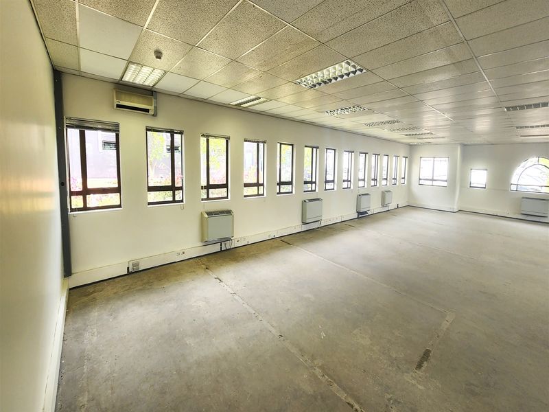 Spacious, empty office area featuring a suspended ceiling, multiple windows for natural light, and wall-mounted air conditioning units.