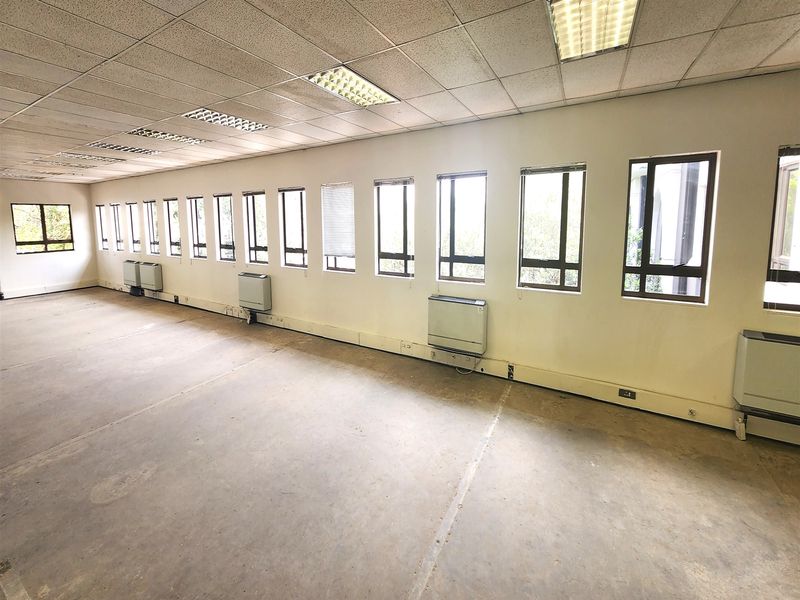 Spacious, empty office area featuring a suspended ceiling, recessed lighting, multiple windows for natural light, and wall-mounted HVAC units.