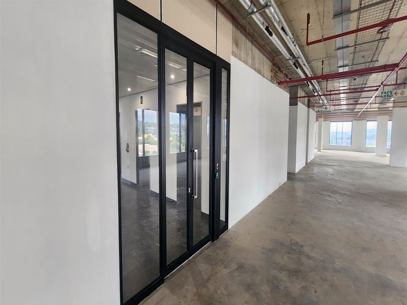 Open-plan office space featuring exposed industrial ceiling services, concrete flooring, and a modern glass-partitioned entrance.