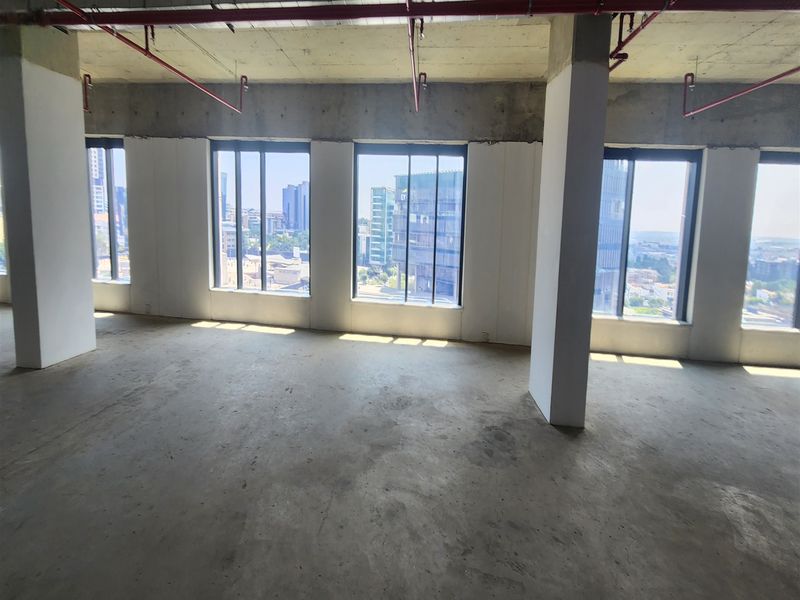 An open-plan office space in shell condition, featuring exposed concrete ceilings, fire sprinkler systems, and large windows with expansive city views.