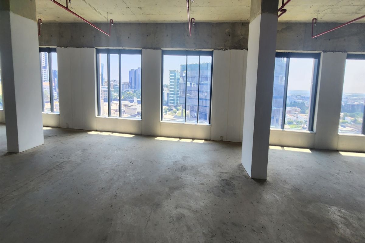 An open-plan office space in shell condition, featuring exposed concrete ceilings, fire sprinkler systems, and large windows with expansive city views.