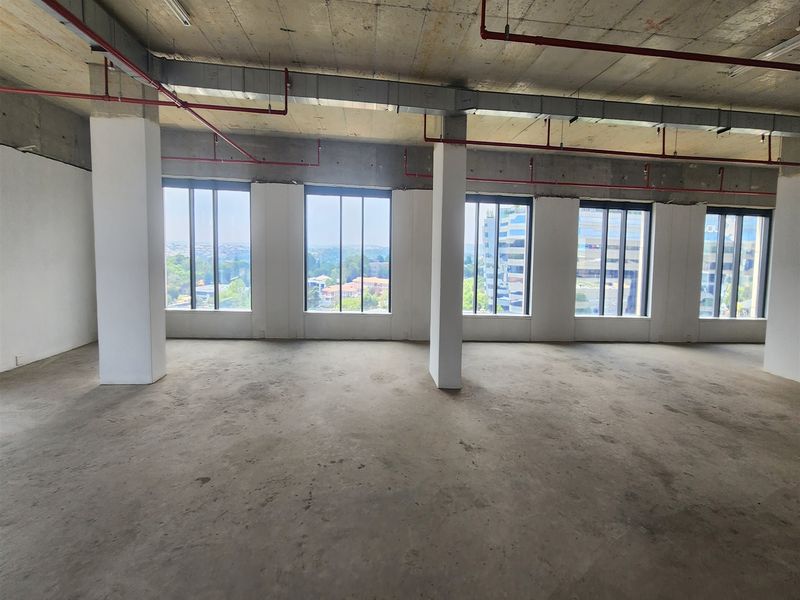 An open-plan office space in shell condition, featuring exposed industrial ceilings, structural columns, and large windows offering expansive city views.
