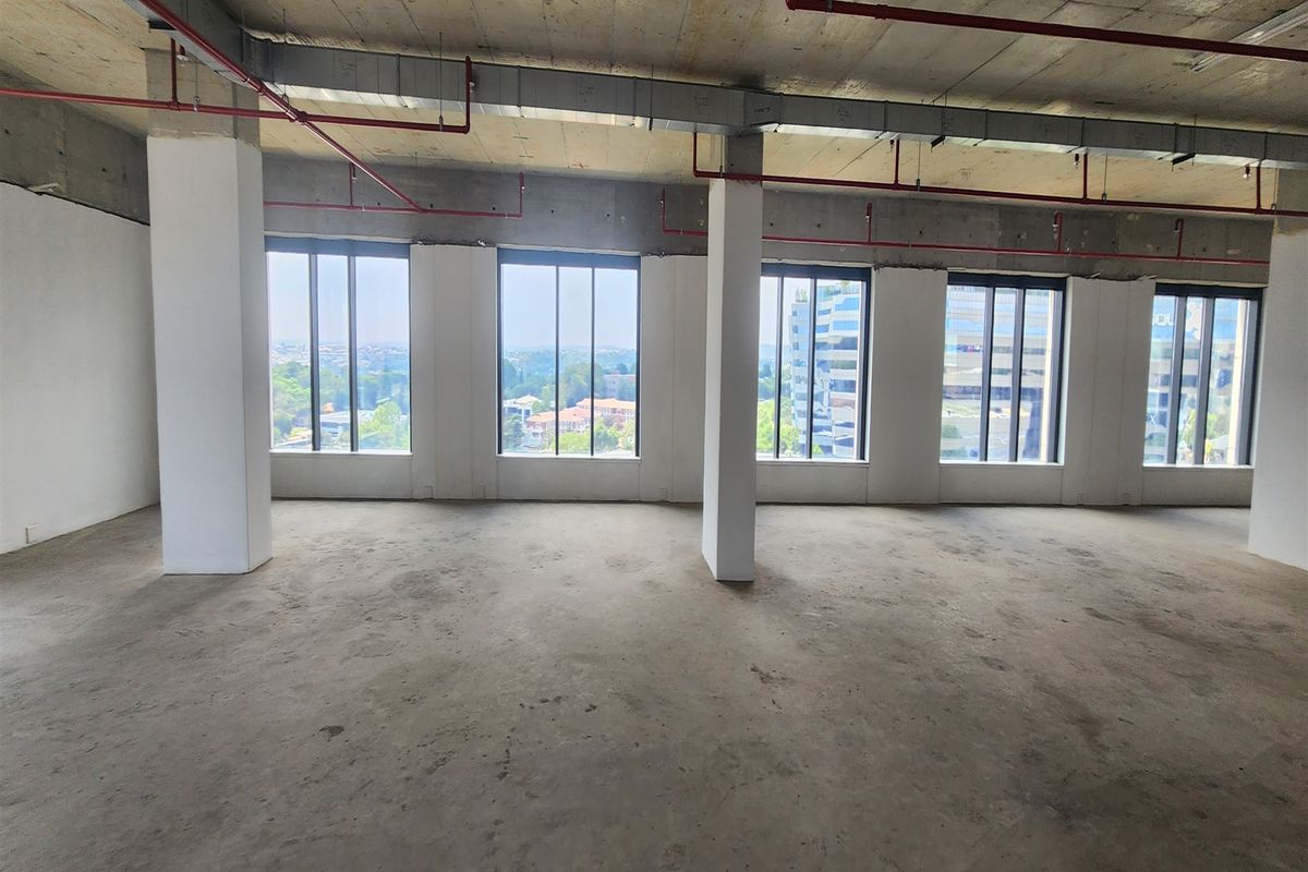 An open-plan office space in shell condition, featuring exposed industrial ceilings, structural columns, and large windows offering expansive city views.