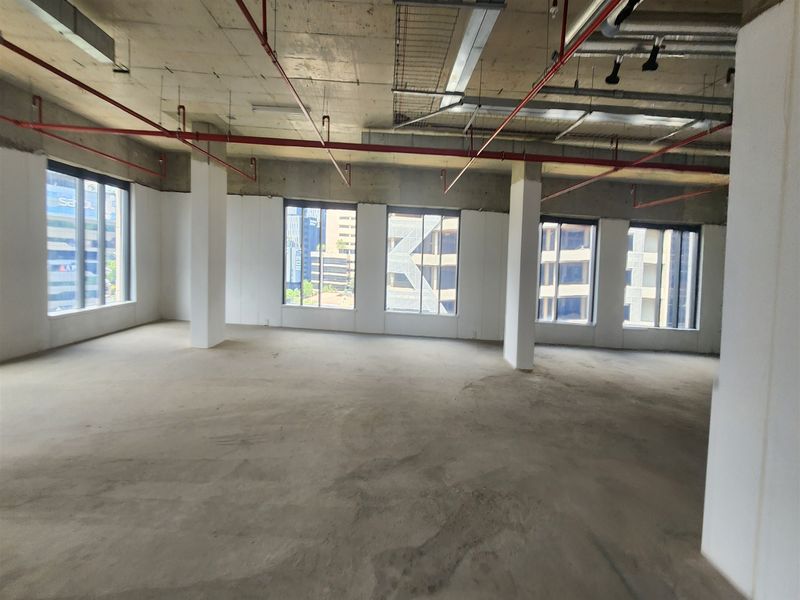 An open-plan office space in shell condition featuring exposed concrete ceilings, fire sprinkler systems, and large windows providing ample natural light.