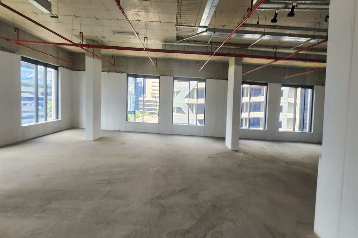 An open-plan office space in shell condition featuring exposed concrete ceilings, fire sprinkler systems, and large windows providing ample natural light.