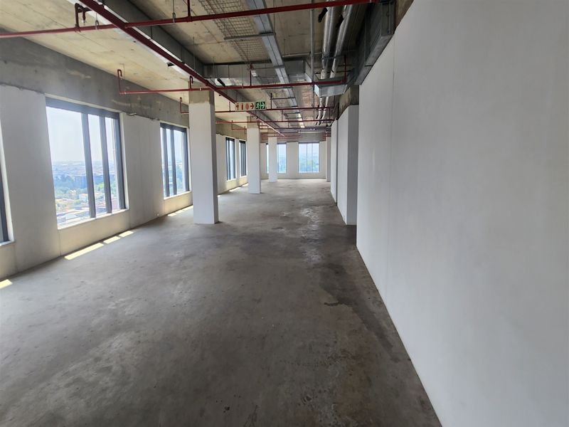 An expansive, open-plan office space in shell condition, featuring exposed industrial ceilings, concrete flooring, and large windows providing ample natural light.
