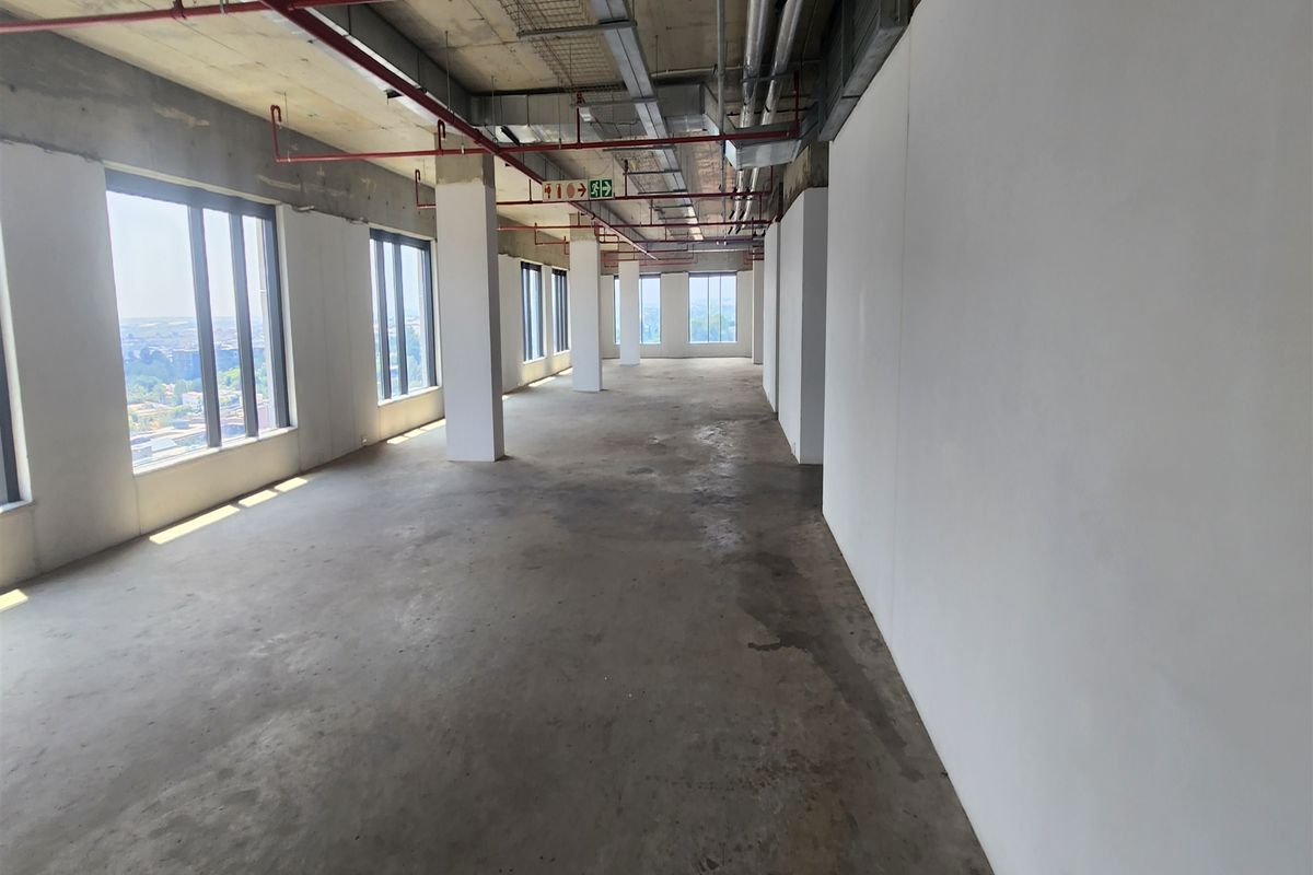 An expansive, open-plan office space in shell condition, featuring exposed industrial ceilings, concrete flooring, and large windows providing ample natural light.