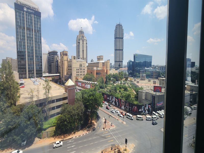 An elevated urban view overlooking the Sandton city skyline, featuring prominent high-rise buildings, surrounding infrastructure, and busy street-level traffic.