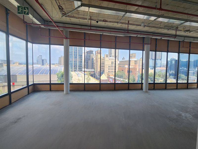 An expansive, unfinished open-plan office space featuring floor-to-ceiling windows with panoramic city views and exposed industrial ceiling infrastructure.