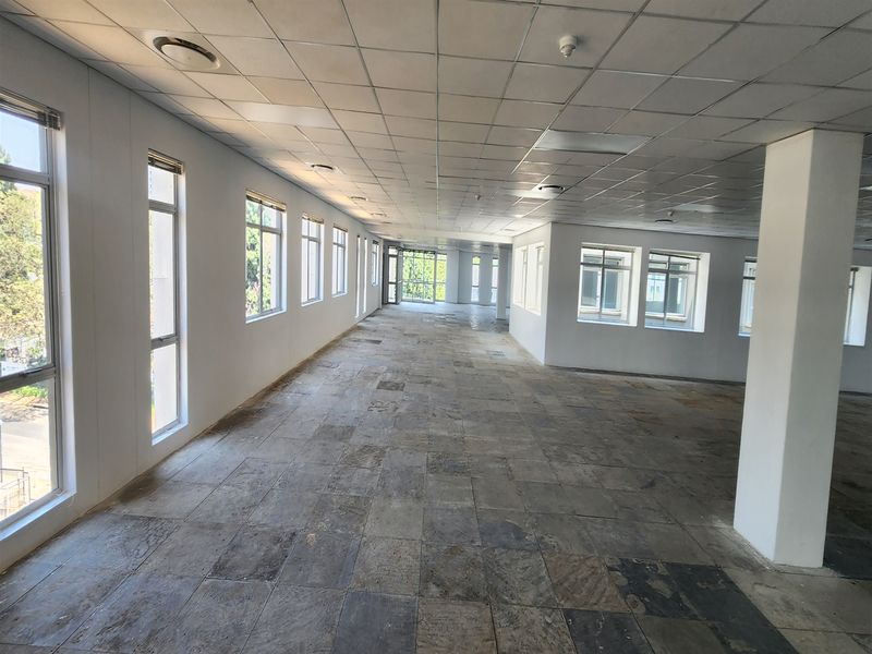 A spacious, open-plan office area featuring large windows, a suspended ceiling, and worn stone tile flooring, offering a blank canvas for customization.