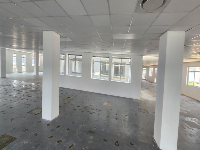 An expansive, open-plan office space featuring a suspended ceiling, structural columns, and large windows, currently requiring flooring repairs.