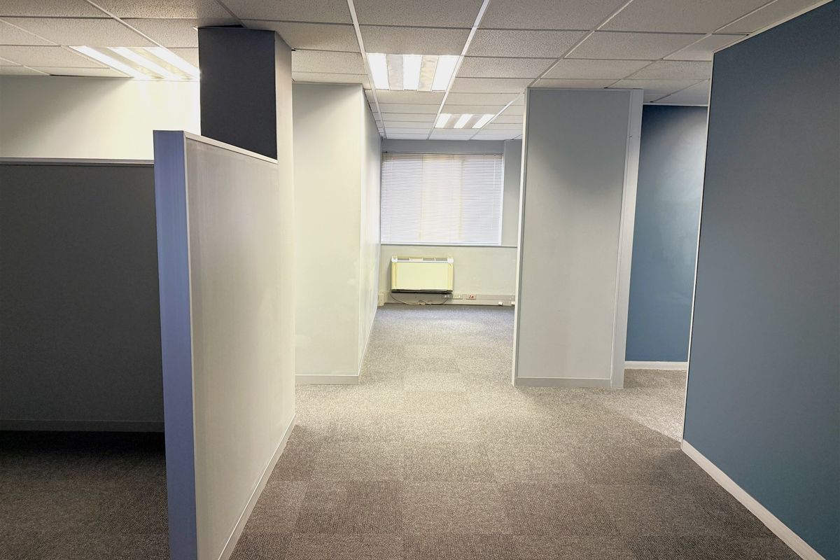 An empty office space featuring carpet tiles, suspended ceilings with recessed lighting, partition walls, and a wall-mounted HVAC unit.