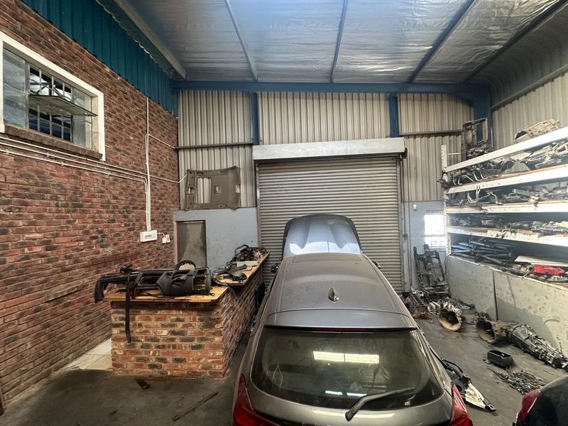 A commercial garage space featuring exposed brick walls, a roller shutter door, and industrial shelving, currently utilized for automotive storage and parts.