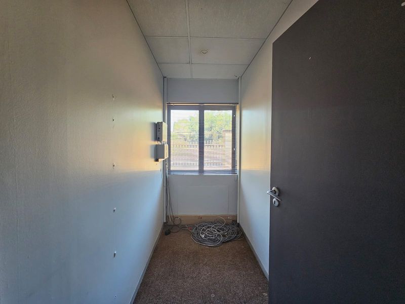 A small, empty office space featuring carpeted flooring, a suspended ceiling, and a window with blinds, currently requiring minor cosmetic repairs.