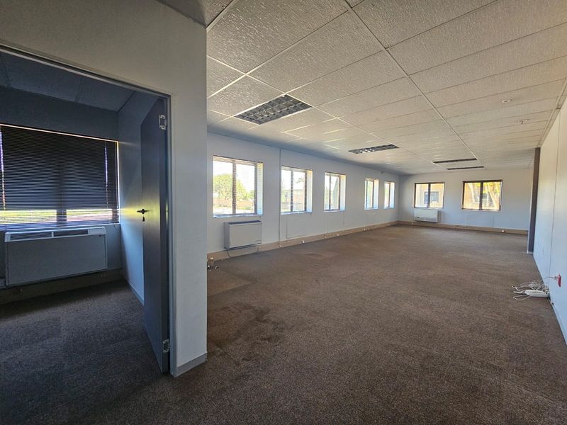 Spacious open-plan office area featuring suspended ceilings, recessed lighting, wall-mounted HVAC units, and large windows providing ample natural light.
