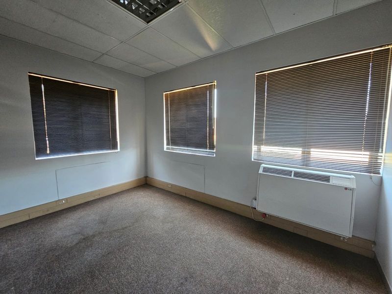 An empty office space featuring carpet flooring, suspended ceiling, and a wall-mounted air conditioning unit with venetian blinds on the windows.