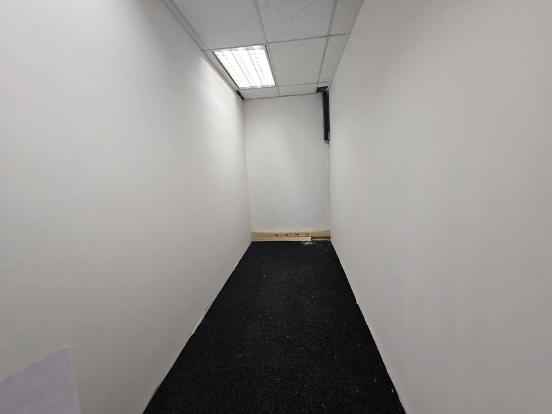 A small, empty office space featuring a suspended ceiling with recessed lighting, dark carpet flooring, and wall-mounted electrical trunking.