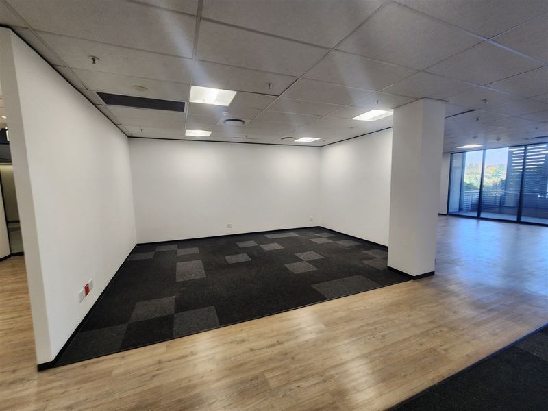 A spacious, empty open-plan office area featuring a mix of carpet and laminate flooring, suspended ceilings, and large windows providing natural light.