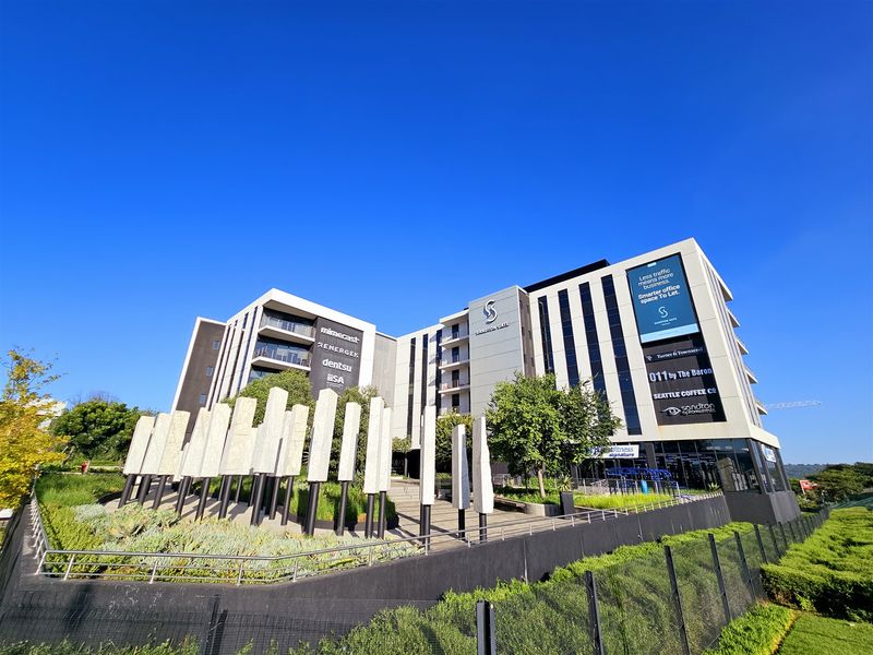 A modern commercial office building facade featuring landscaped gardens, concrete architectural pillars, and prominent tenant signage under a clear blue sky.