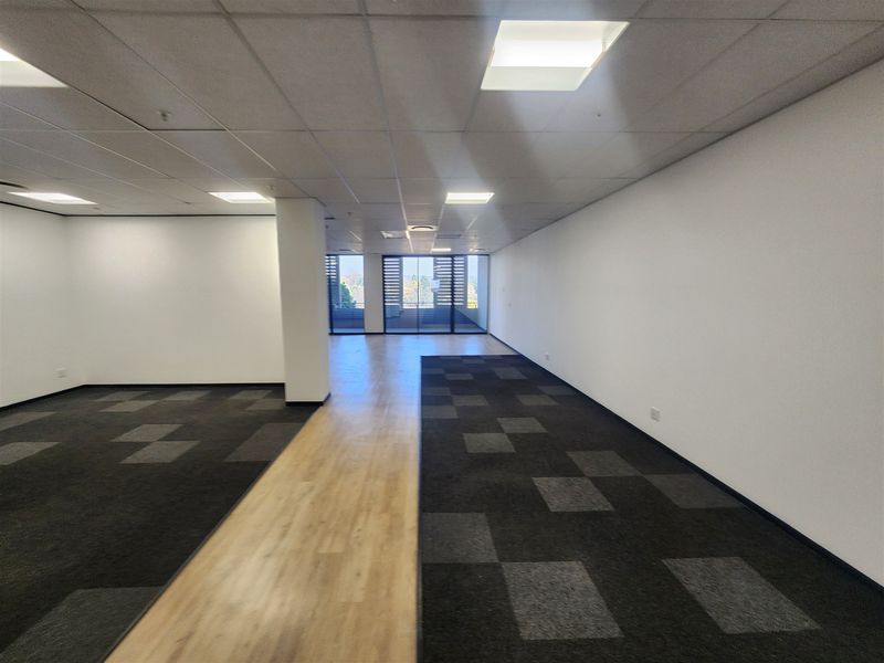 A spacious, empty open-plan office area featuring a mix of carpet and laminate flooring, recessed ceiling lights, and large windows providing natural light.
