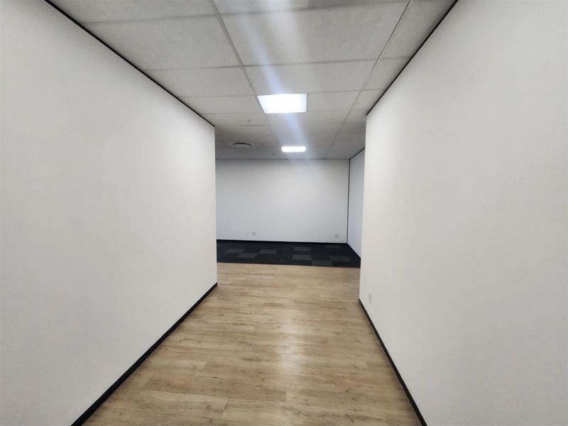 A clean, vacant office space featuring a mix of laminate and carpet flooring, suspended ceilings with recessed lighting, and neutral white walls.