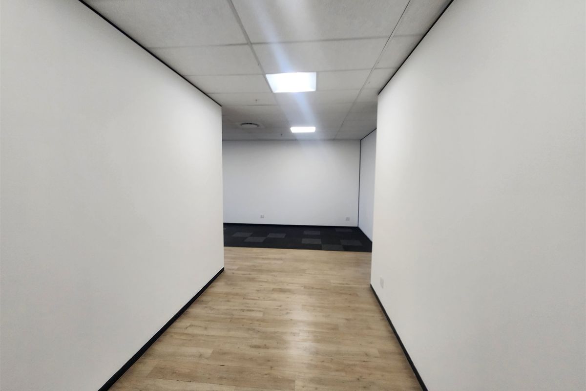 A clean, vacant office space featuring a mix of laminate and carpet flooring, suspended ceilings with recessed lighting, and neutral white walls.