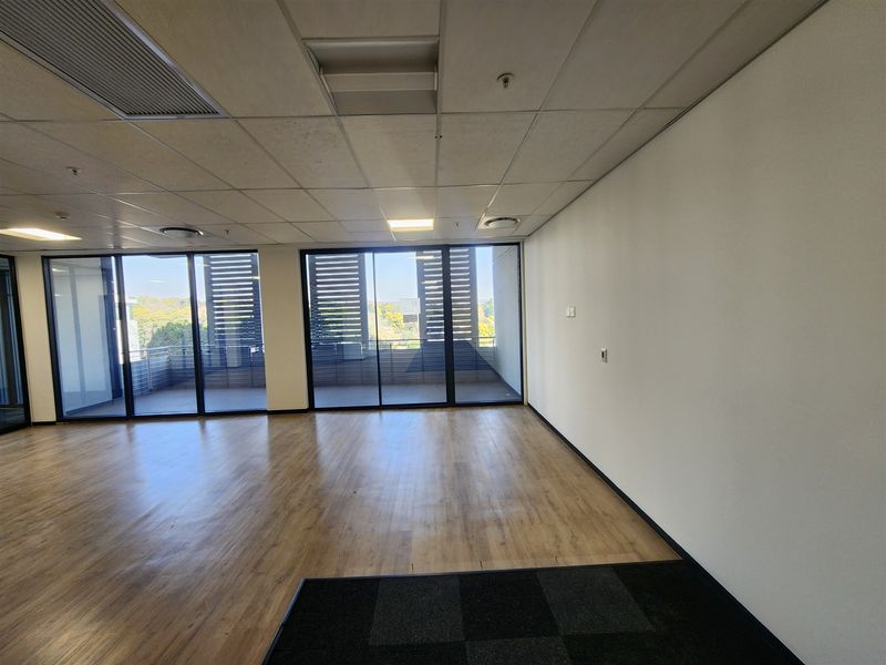 A bright, empty office space featuring wood-look laminate flooring, carpeted sections, suspended ceilings, and large sliding glass doors leading to an outdoor balcony.