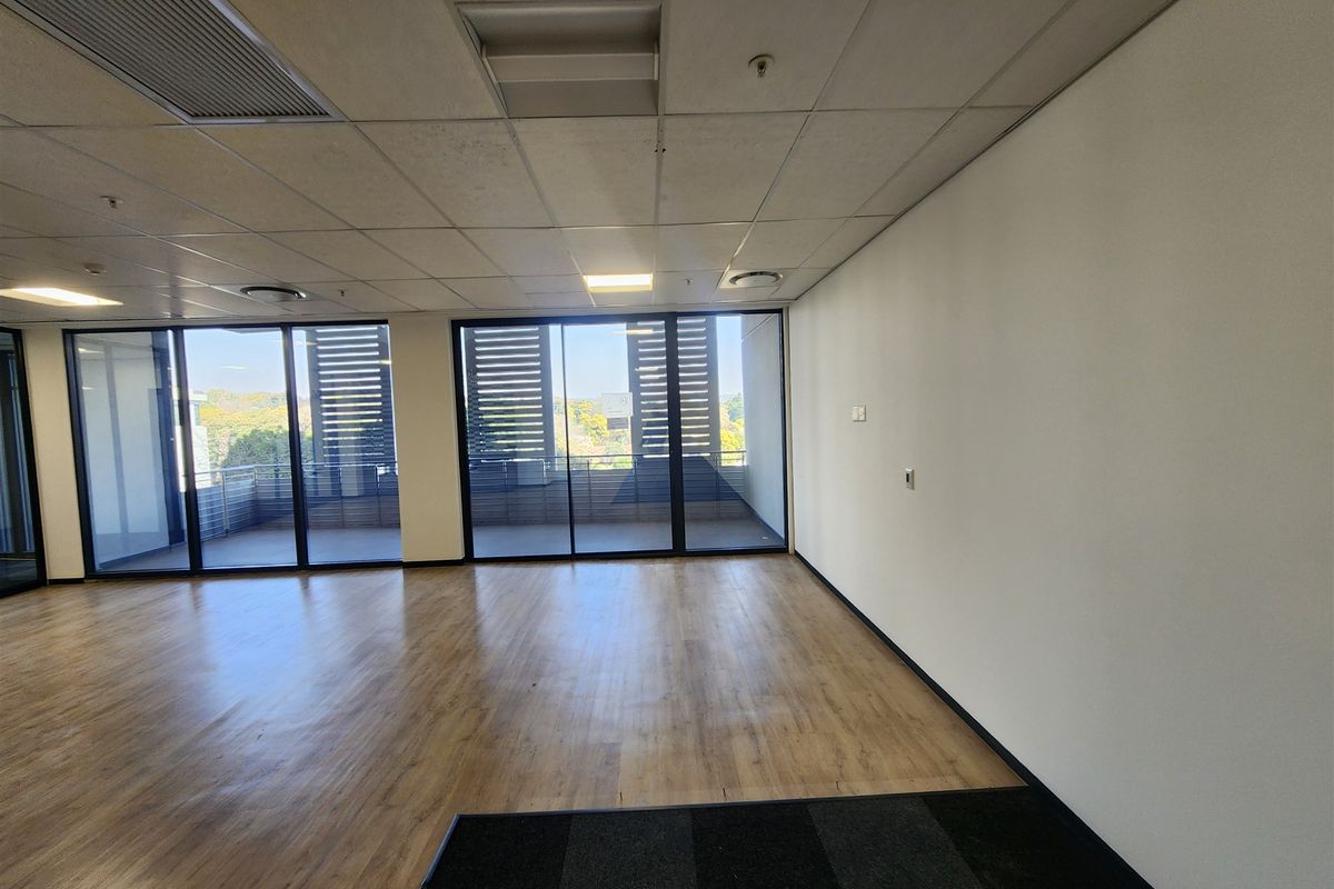 A bright, empty office space featuring wood-look laminate flooring, carpeted sections, suspended ceilings, and large sliding glass doors leading to an outdoor balcony.