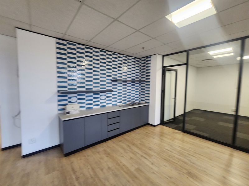 Modern office space featuring a built-in kitchenette with blue and white tiled backsplash, glass partition walls, and a mix of laminate and carpet flooring.
