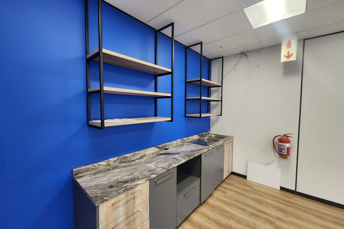 A compact office kitchenette featuring modern grey and wood-grain cabinetry, marble-effect countertops, open metal shelving, and a blue accent wall.