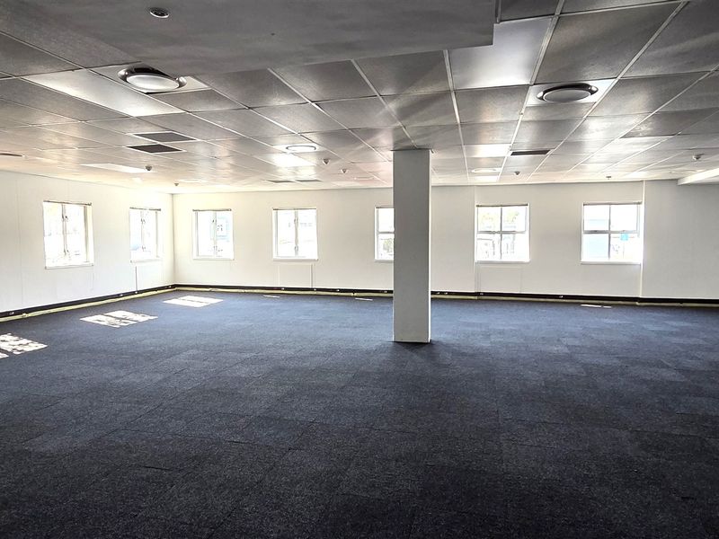 A spacious, open-plan office area featuring a suspended ceiling, recessed lighting, and dark carpet tiles, providing a versatile workspace.